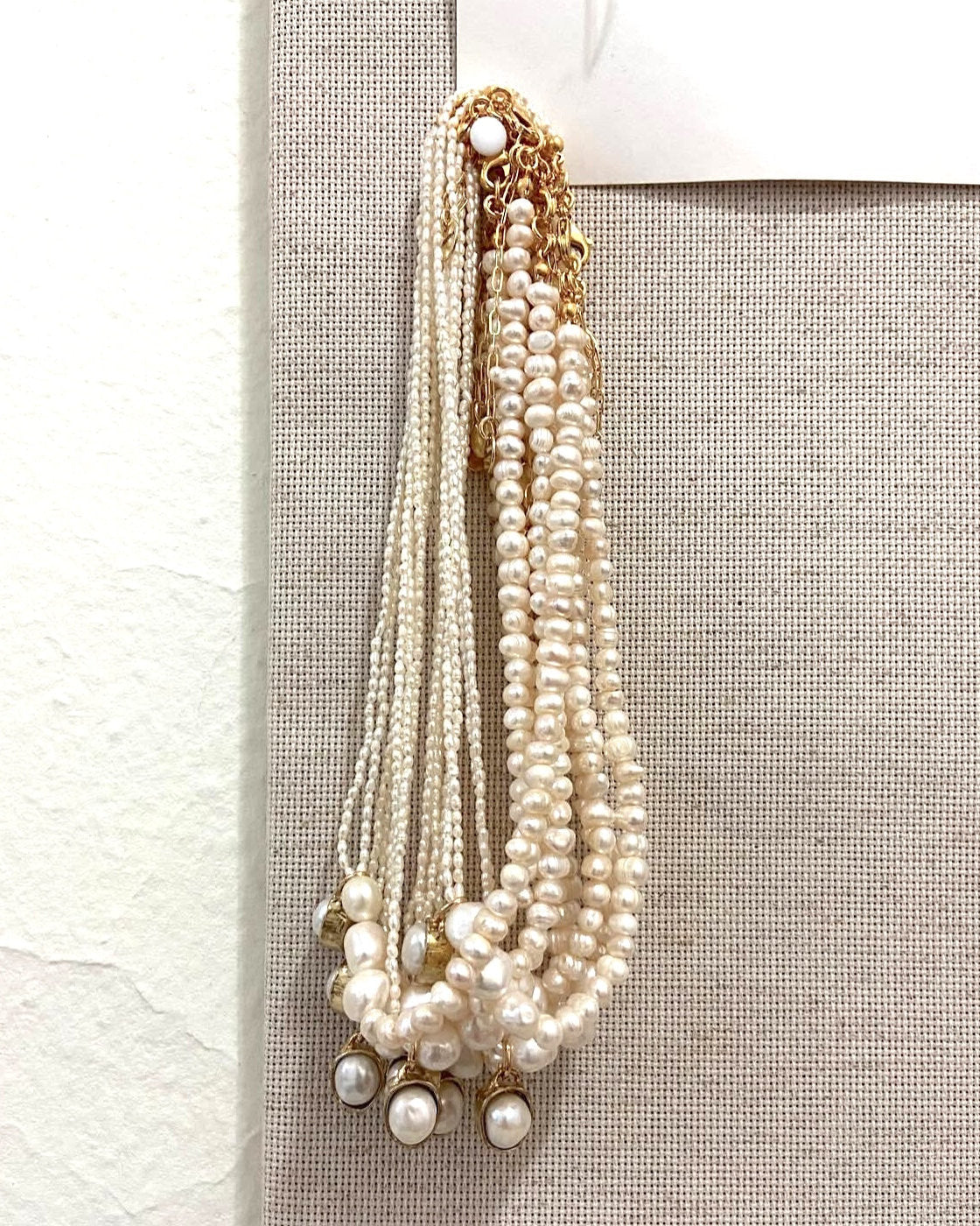 A multi-strand pearl necklace with gold chain details and pearl drop charms, displayed on a textured beige fabric jewelry stand.