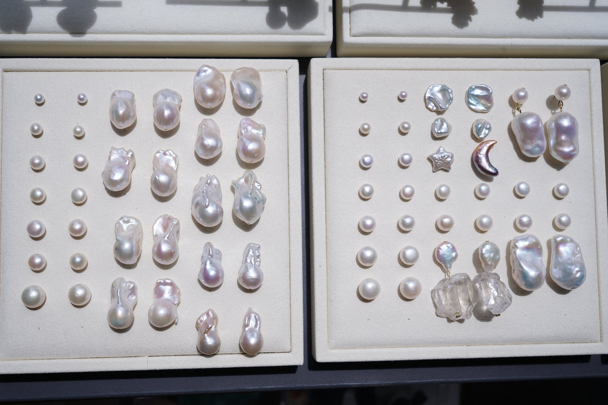 Two cream-colored display trays holding assorted baroque pearls and pearl-like beads in various sizes and shapes, arranged in rows.