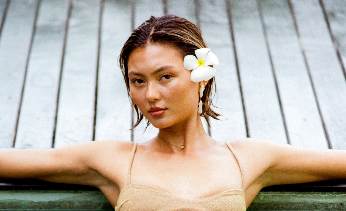A woman with a white plumeria flower in her hair leans against the edge of a pool wearing a beige cutout bikini and dangling pearl earrings. Her reflection is visible in the water.