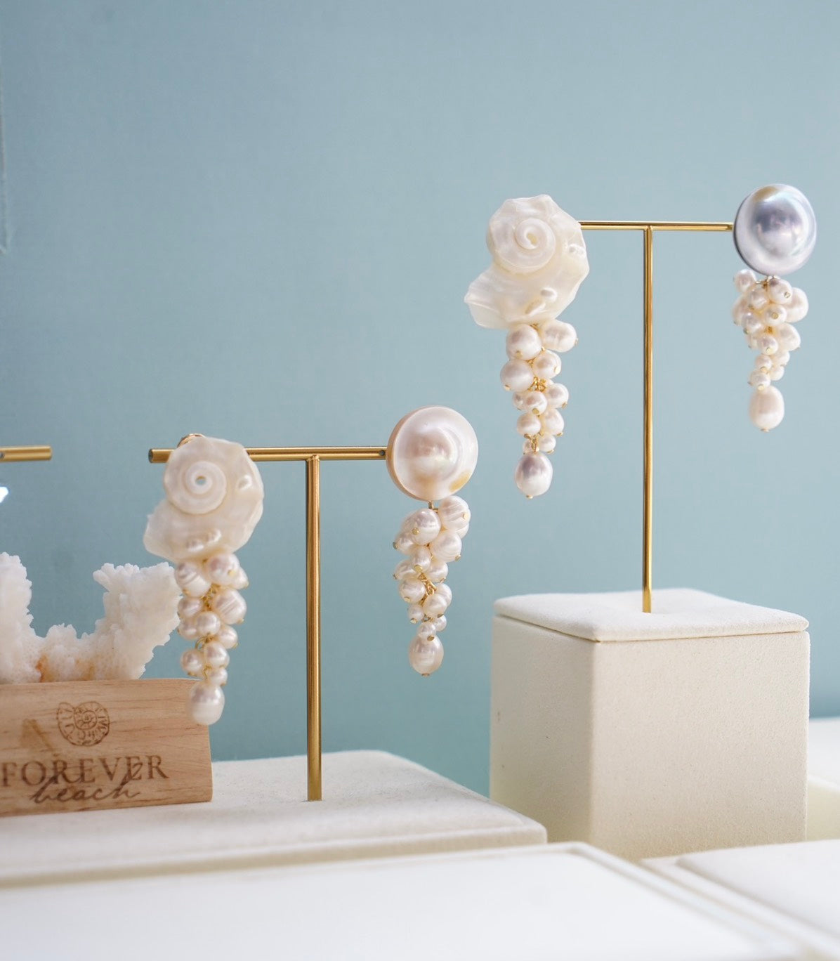 Elegant dangle earrings displayed on gold jewelry stands, featuring white and iridescent pearls arranged in floral and cascading clusters, set against a soft light blue background.