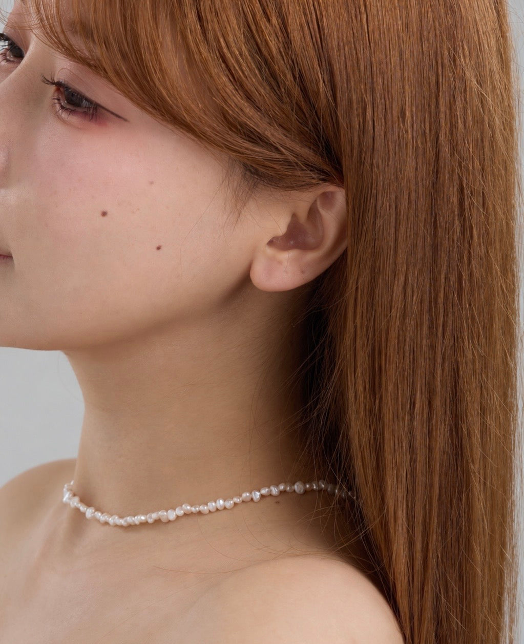 A close-up side profile of a woman with long auburn hair wearing a delicate pearl choker necklace against a light gray background.