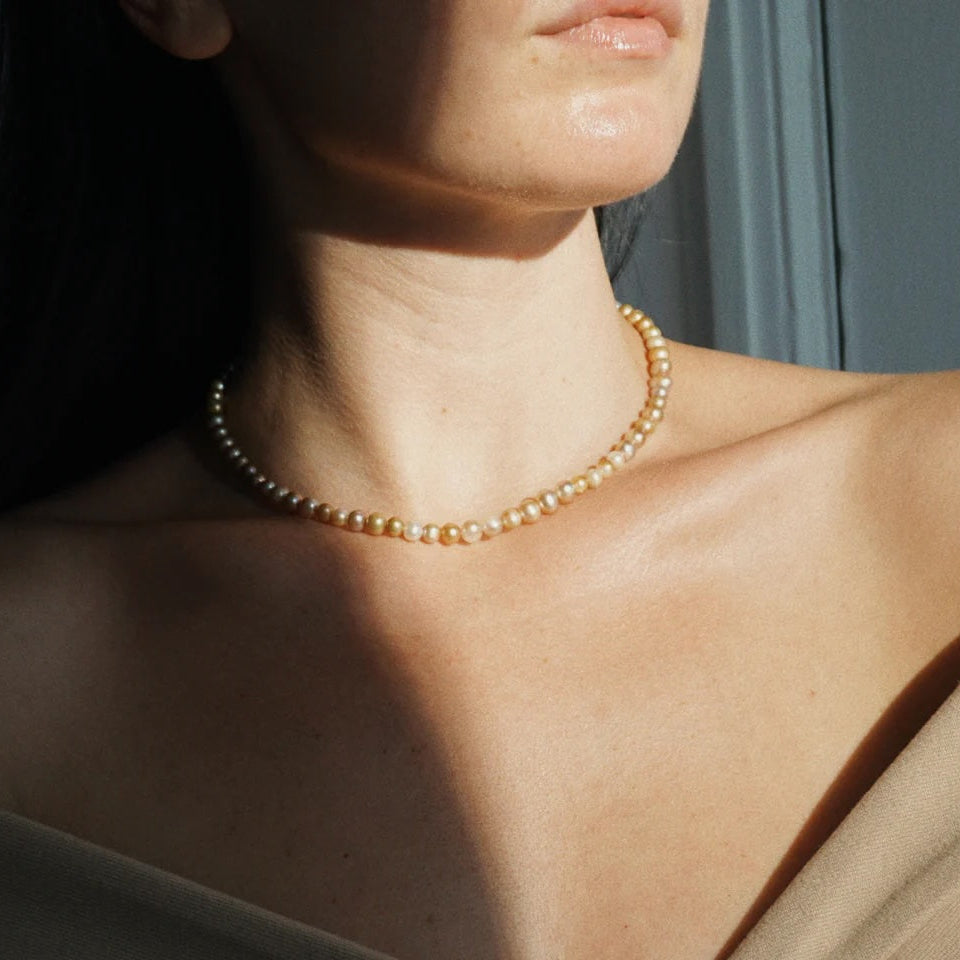 A close-up lifestyle shot of a model wearing a delicate strand of cream-colored freshwater pearls as a choker necklace, styled with a beige off-the-shoulder top under warm, direct sunlight.