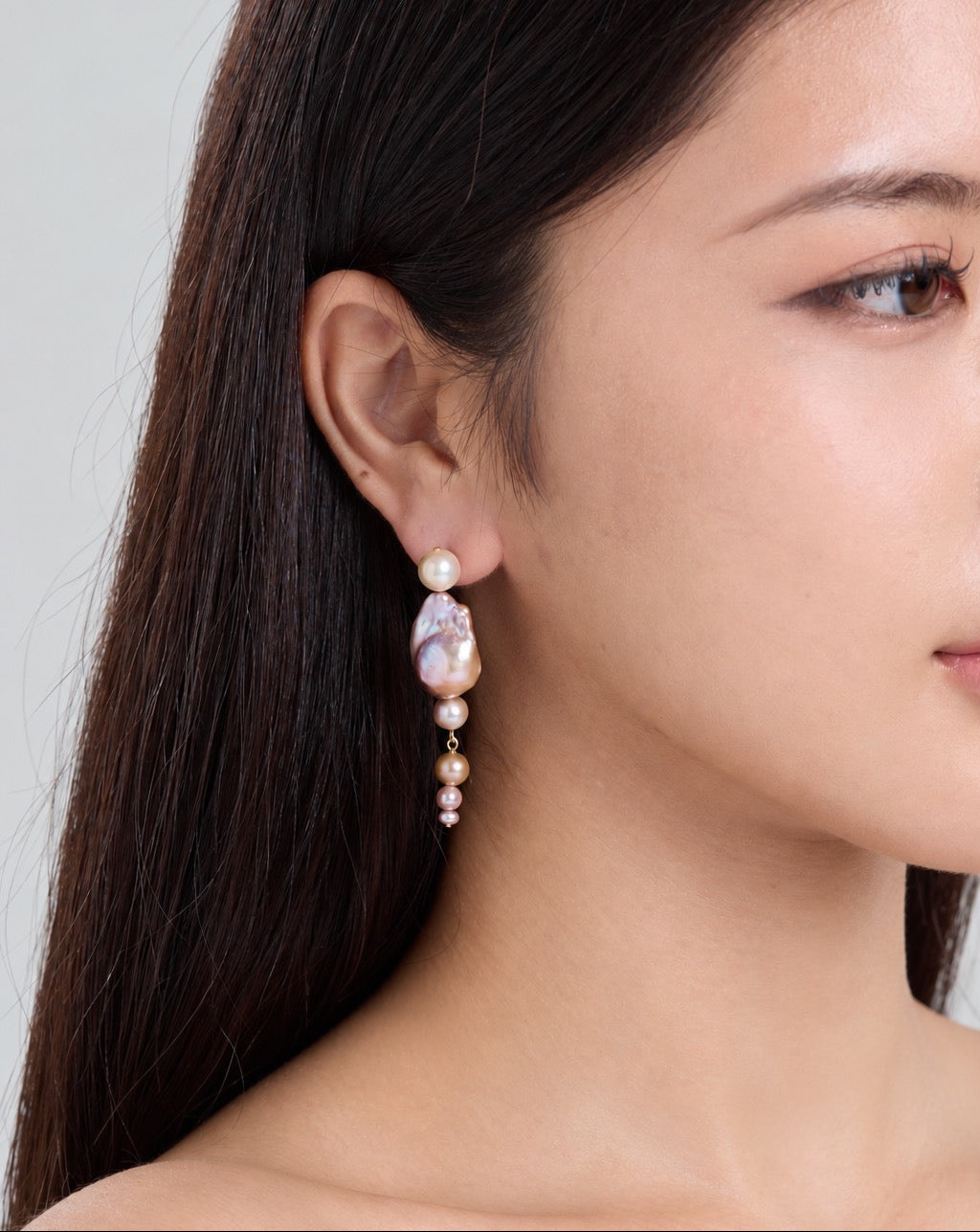 A close-up side profile of a woman wearing a dangling earring. The earring features a small gold-toned bead, a white pearl, and a larger irregular lavender-purple pearls with smaller pearl beads below, set against a neutral gray background.
