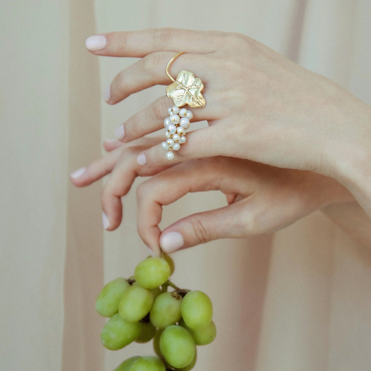A close-up of a hand holding an earring with a gold vine with a pearl grape cluster, and the other hand holding a green grapes against a beige curtain background.