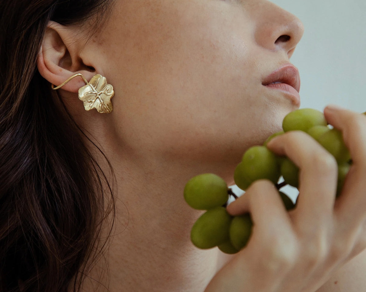 A close-up lifestyle shot of a model wearing a golden earring shaped like a textured leaf. The model is holding a bunch of green grapes against a soft, light gray background.