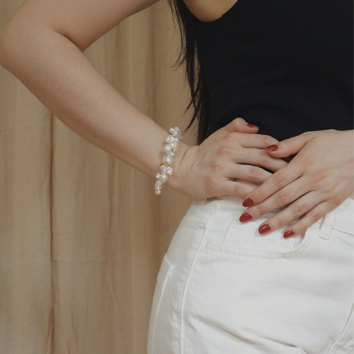 A close-up of a person wearing a pearl bracelet on their left wrist, paired with a black top and white pants, standing against a beige curtain background.