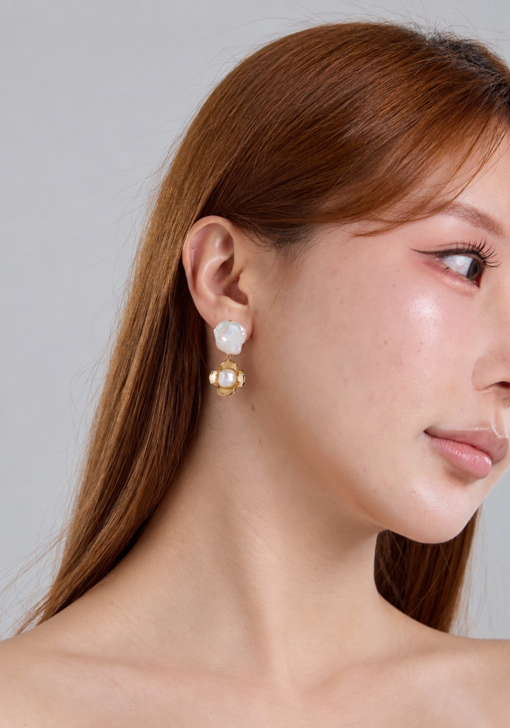 A close-up side profile of a woman with long auburn hair wearing a gold and white dangle earring against a light gray background.