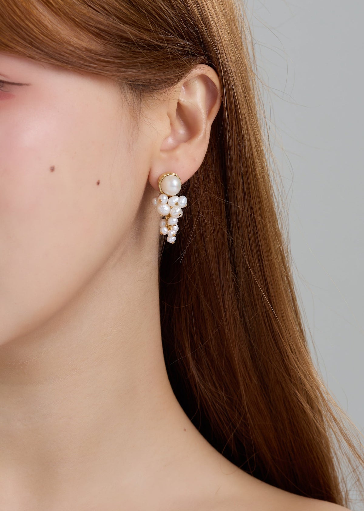 A close-up of a woman's ear wearing a dangling pearl cluster earring with a gold-toned top and small pearl accents, against a soft gray background.