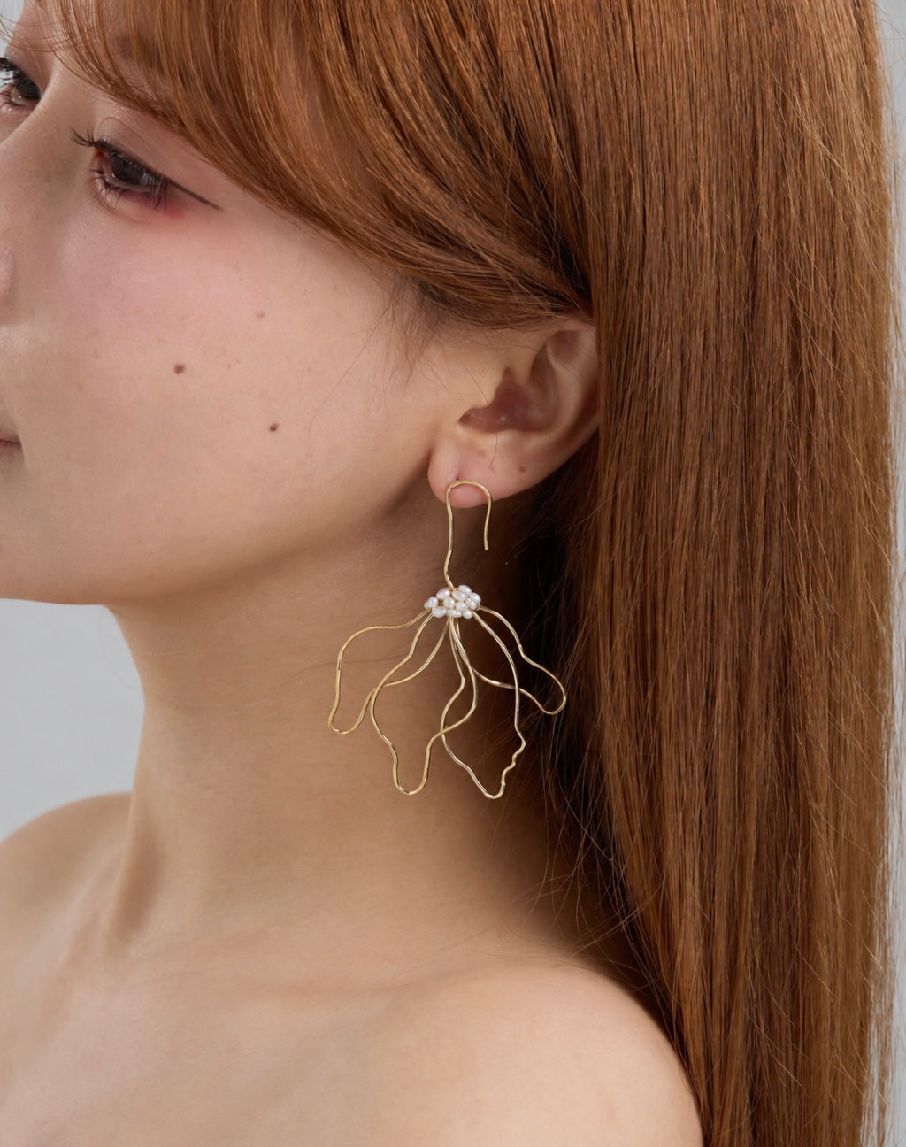A close-up side profile of a woman with long auburn hair wearing a delicate gold wire earring with a small white floral pearl cluster at the top.
