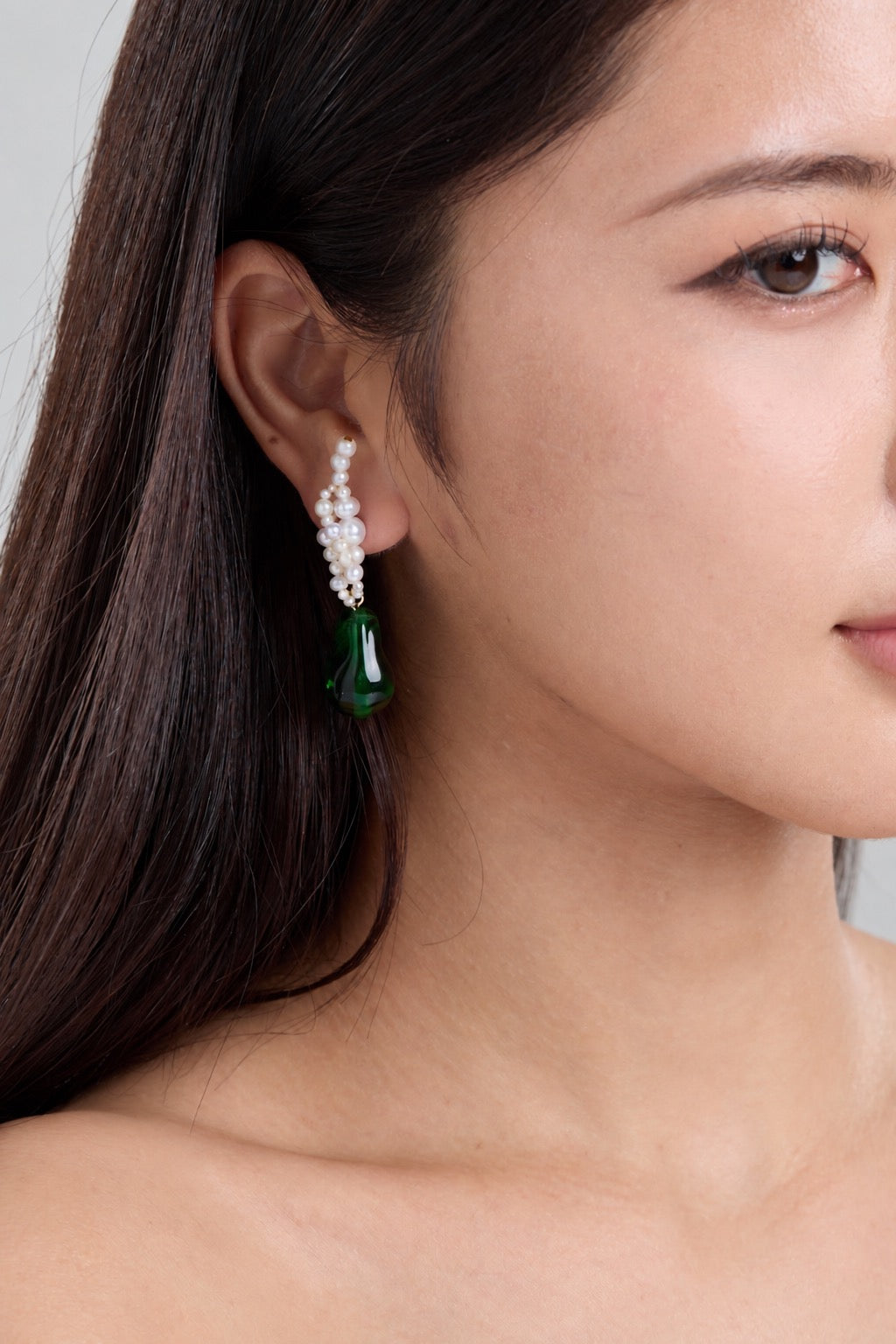 A close-up of a woman's ear wearing a dangling earring with a white pearl cluster top and a green teardrop-shaped gemstone, against a light gray background.