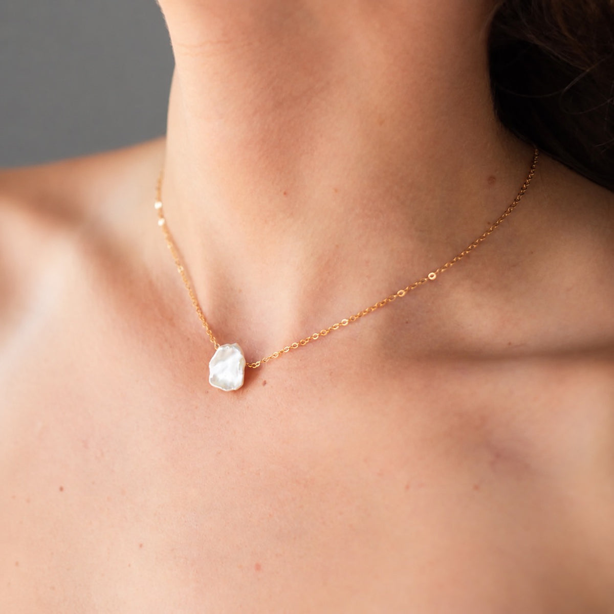 A close-up of a model's neck and chest wearing a delicate gold chain necklace with an organic irregular white pearl pendant.