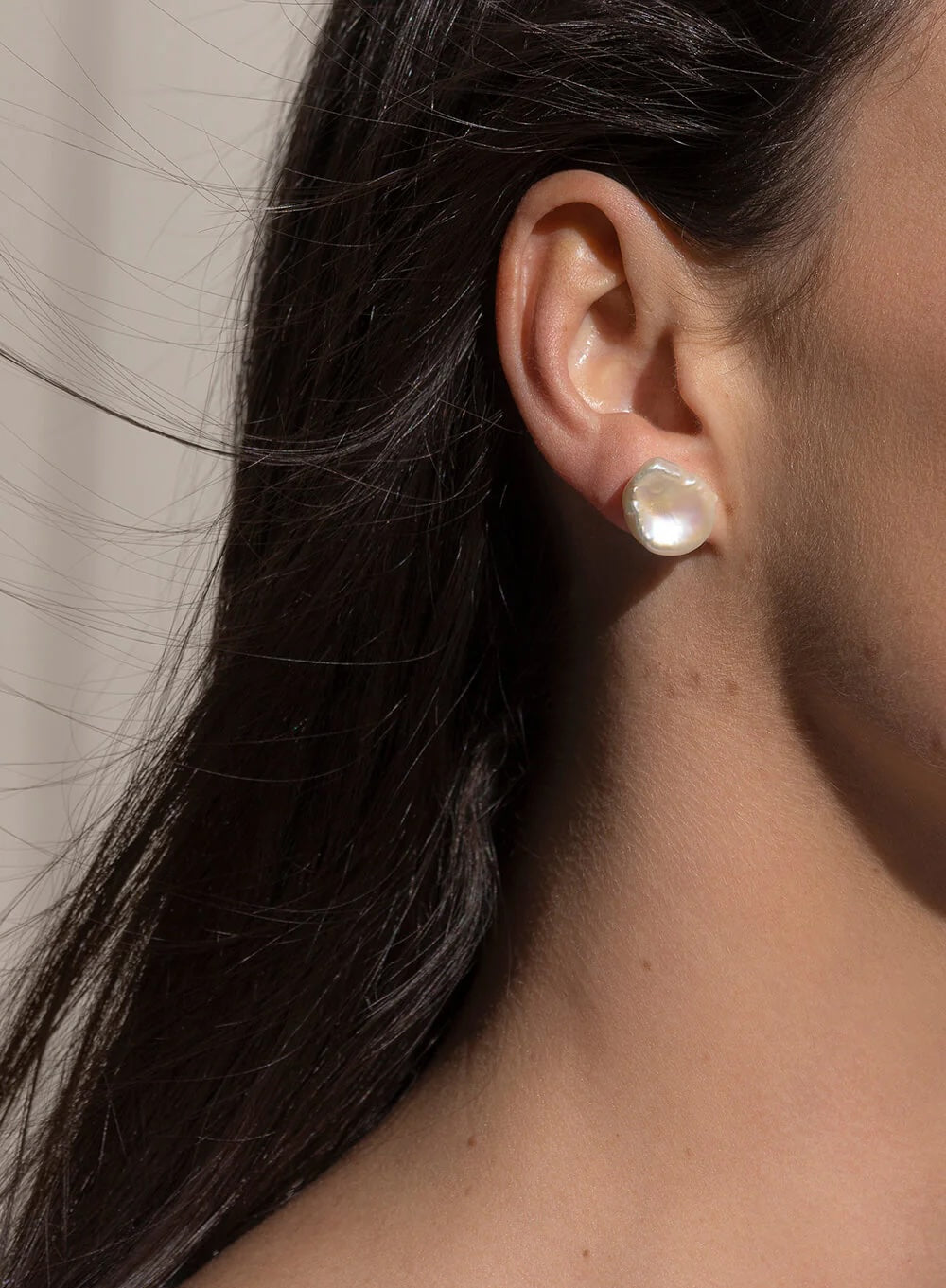 A close-up side profile of a woman's ear wearing a single round, lustrous pearl stud earring with a gold-toned setting.