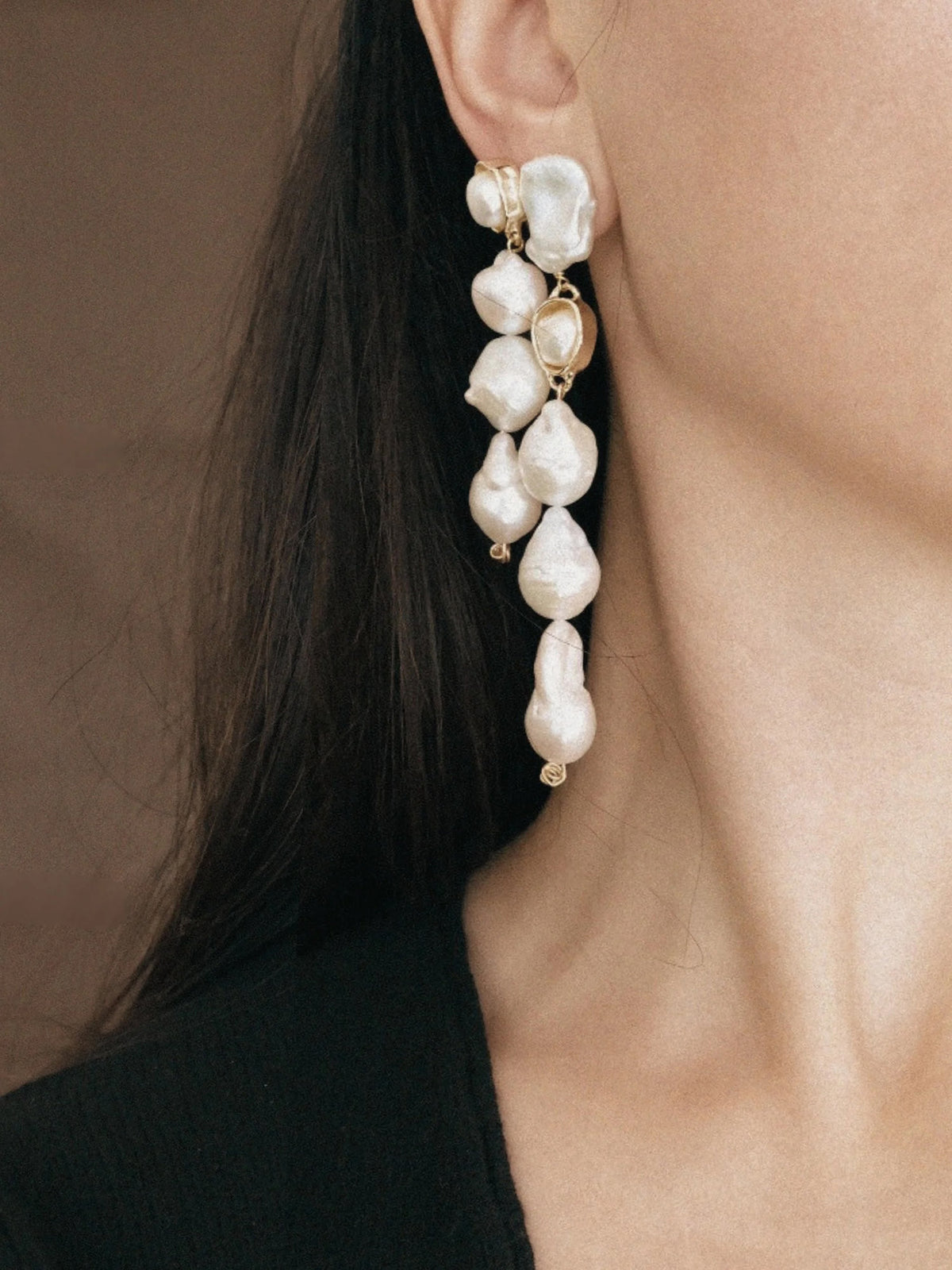 A close-up of a model wearing a long, multi-tiered dangle earring made of irregular white baroque pearls and gold-toned metal settings.