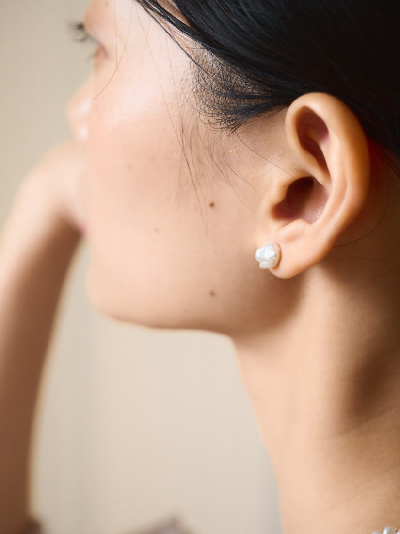 A close-up side profile of a woman wearing a single white pearl stud earring on her earlobe against a soft beige background.