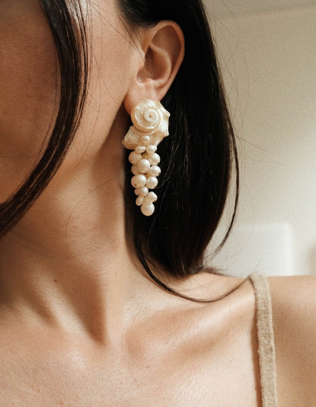 A close-up of a model's ear wearing a dangle earring made of a spiral shell and small pearl cluster, with her dark hair and beige strap visible against a soft neutral background.