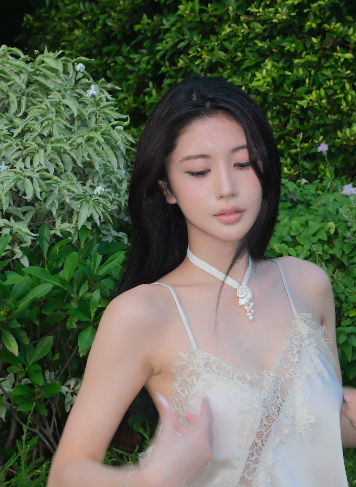A woman with long black hair is wearing a light cream lace camisole top and a matching cream necklace made of a seashell and a pearl cluster, standing outdoors in front of a green garden background.