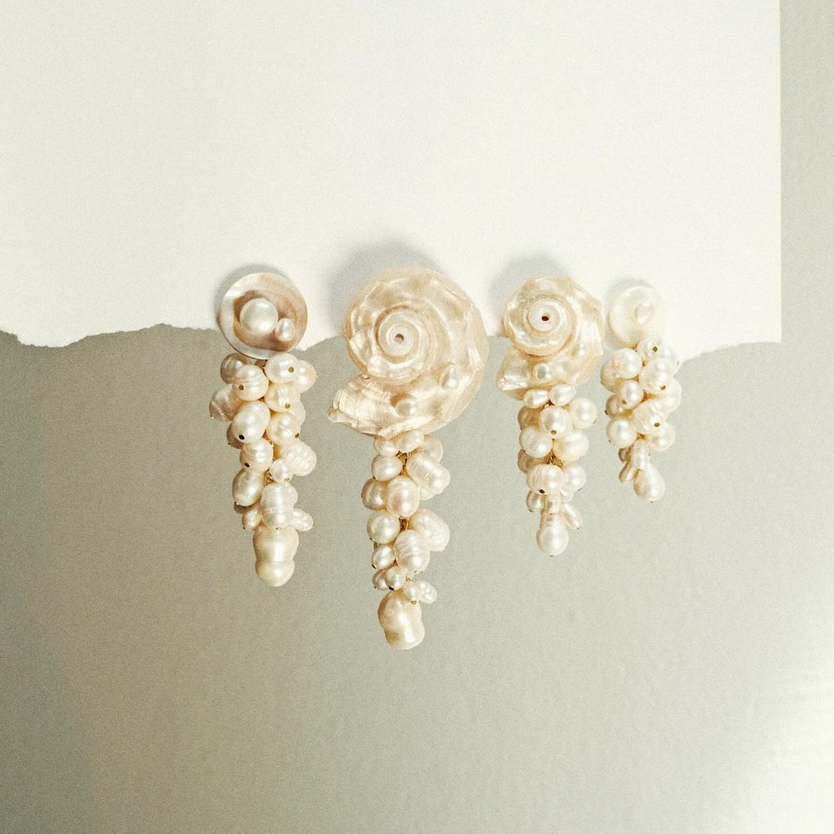 Two pairs of pearl drop earrings with shell details, displayed against a split beige and off-white background.