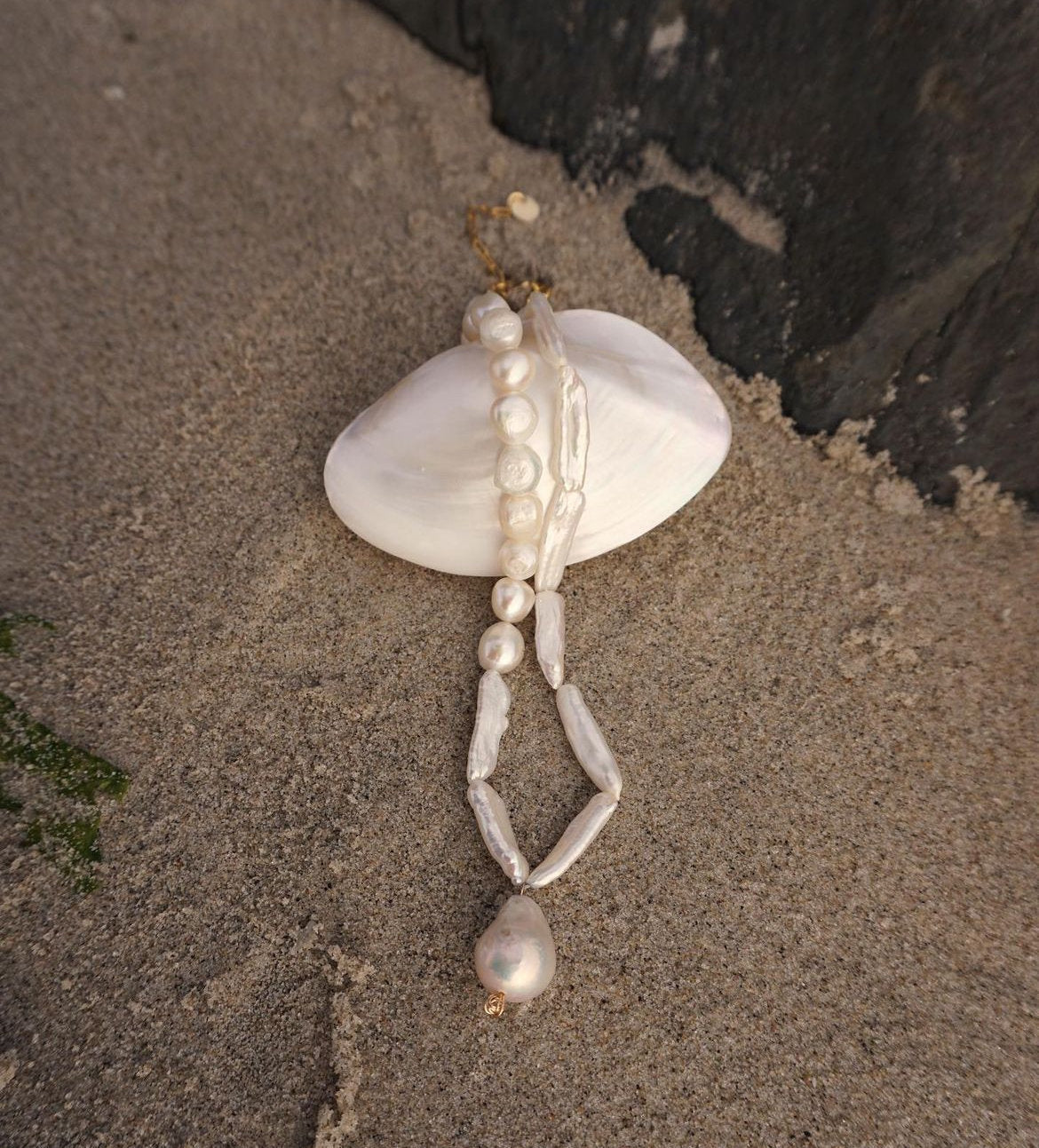 A pearl beaded necklace with irregular white pearls and a gold-toned clasp, laid over a white clam shell on a sandy surface.