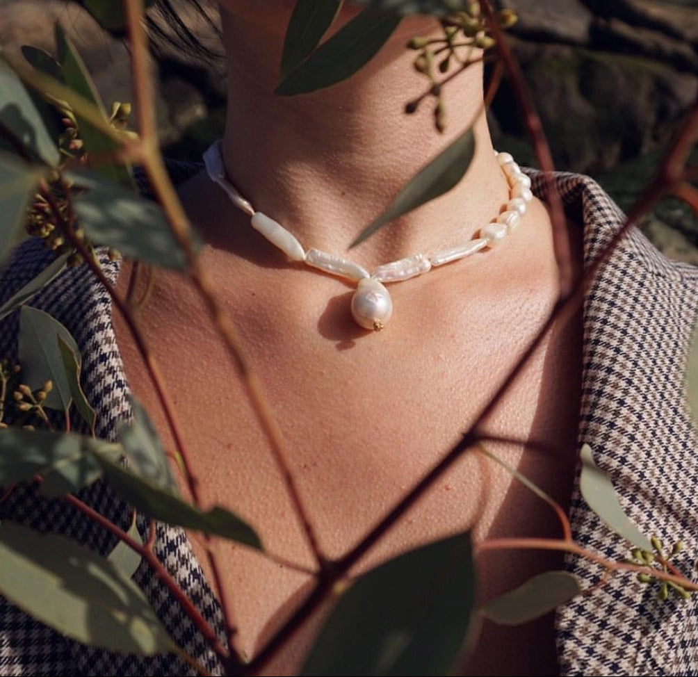 A close-up lifestyle shot of a person wearing a natural pearl necklace with a large teardrop baroque pearl pendant, styled with a checkered shirt and surrounded by eucalyptus leaves.