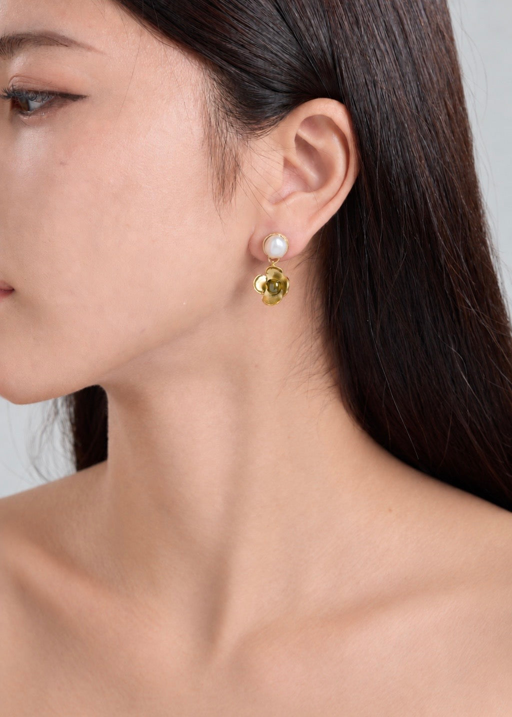 A close-up of a woman's ear wearing a gold flower-shaped stud earring with a round green gemstone in the center and a white pearl at the top.