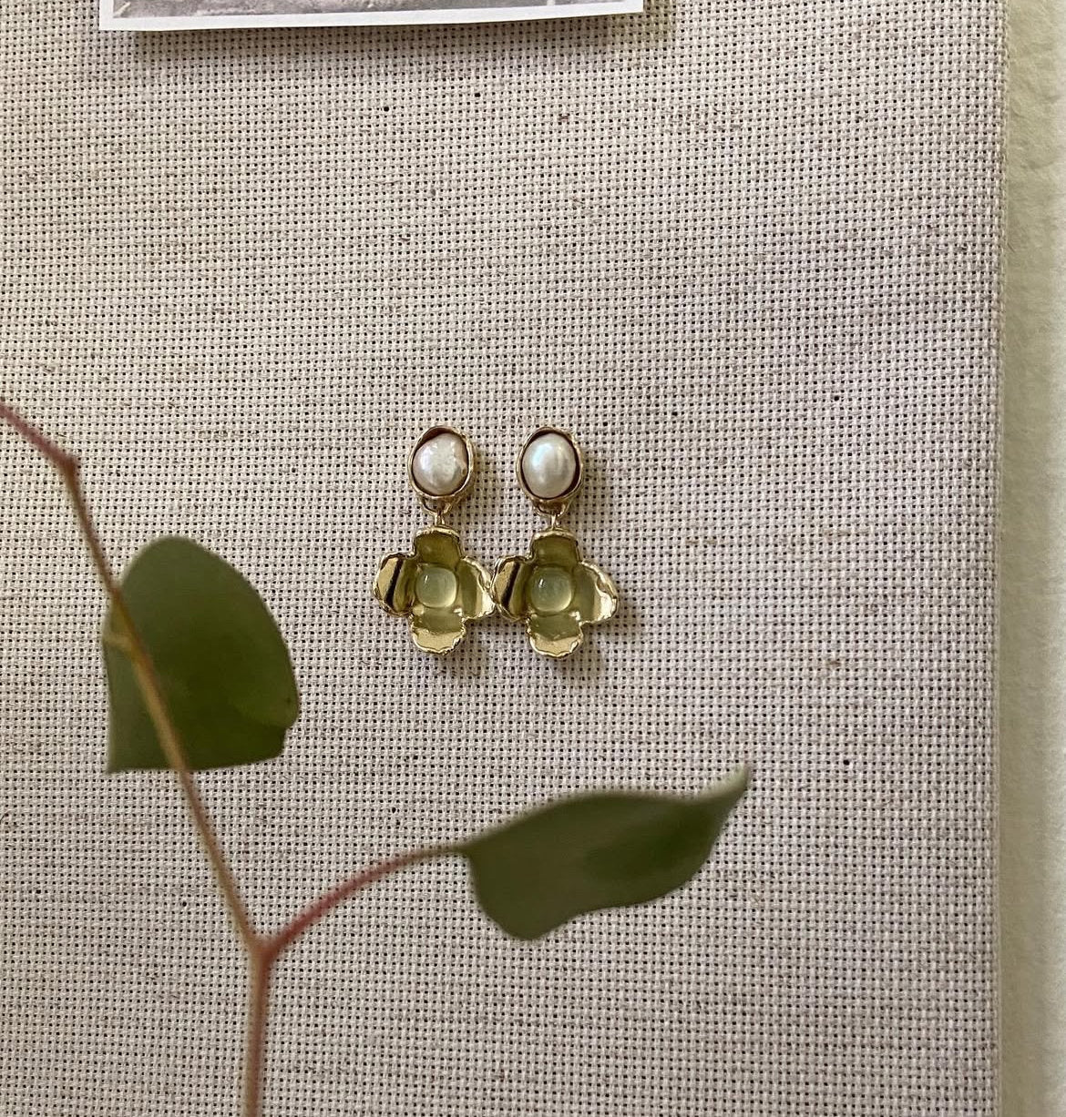 A pair of gold-toned dangle earrings with oval white stones at the top and layered flower-like gold drops below, displayed on a beige woven fabric background with blurred green leaves in the foreground.