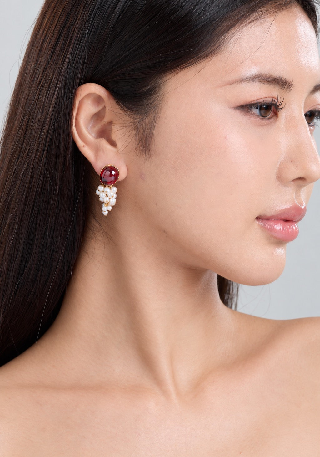 A close-up side profile of a woman wearing a dangling earring with a deep red gemstone and a cluster of small white pearls.