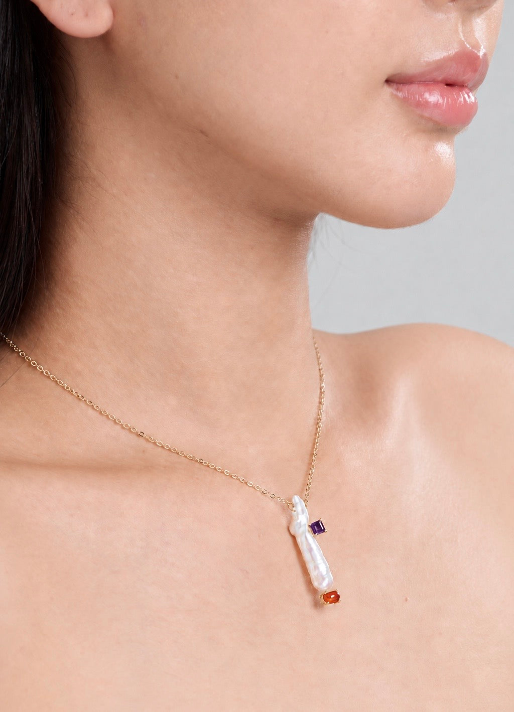 A close-up of a model's neck and upper chest wearing a delicate gold chain necklace with a pendant made from a long white pearl featuring small purple and orange gemstone accents.