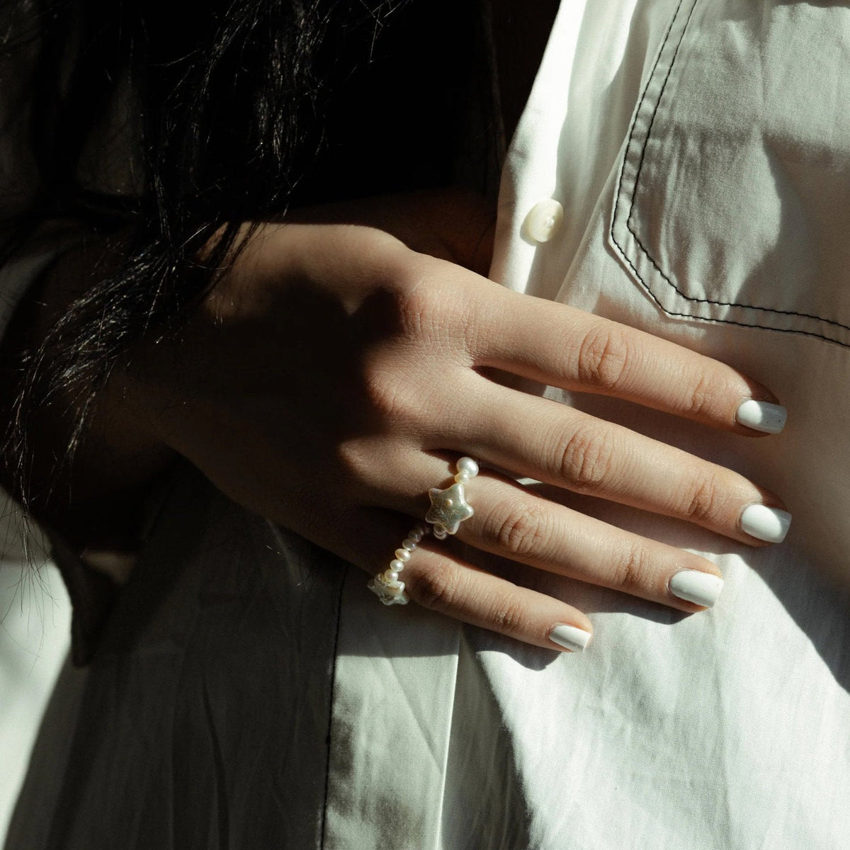 A close-up of a person's hand resting on a white shirt, wearing two delicate pearl rings with star-shaped pearls in the middle