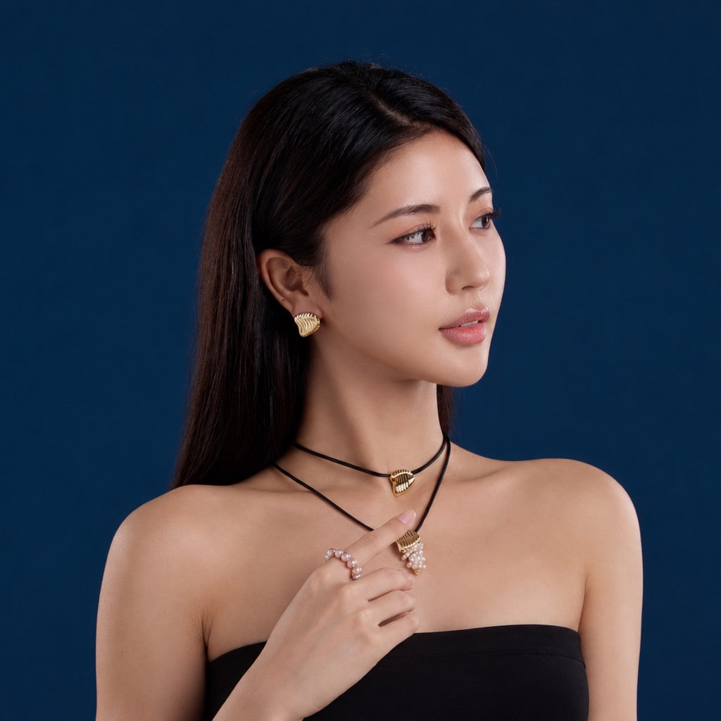 A woman with long dark hair models gold jewelry, including a gold heart-shaped earring, a gold pendant necklace, and rings, against a solid dark blue background.