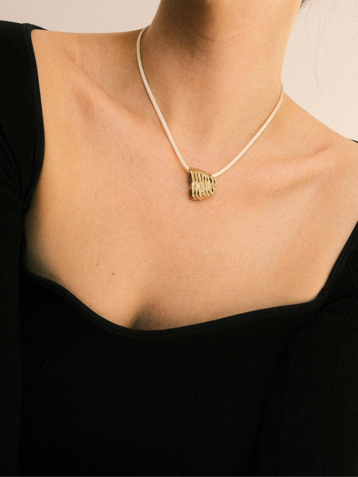 A close-up of a model wearing a delicate white cord necklace with a textured gold pendant, paired with a black ribbed top.