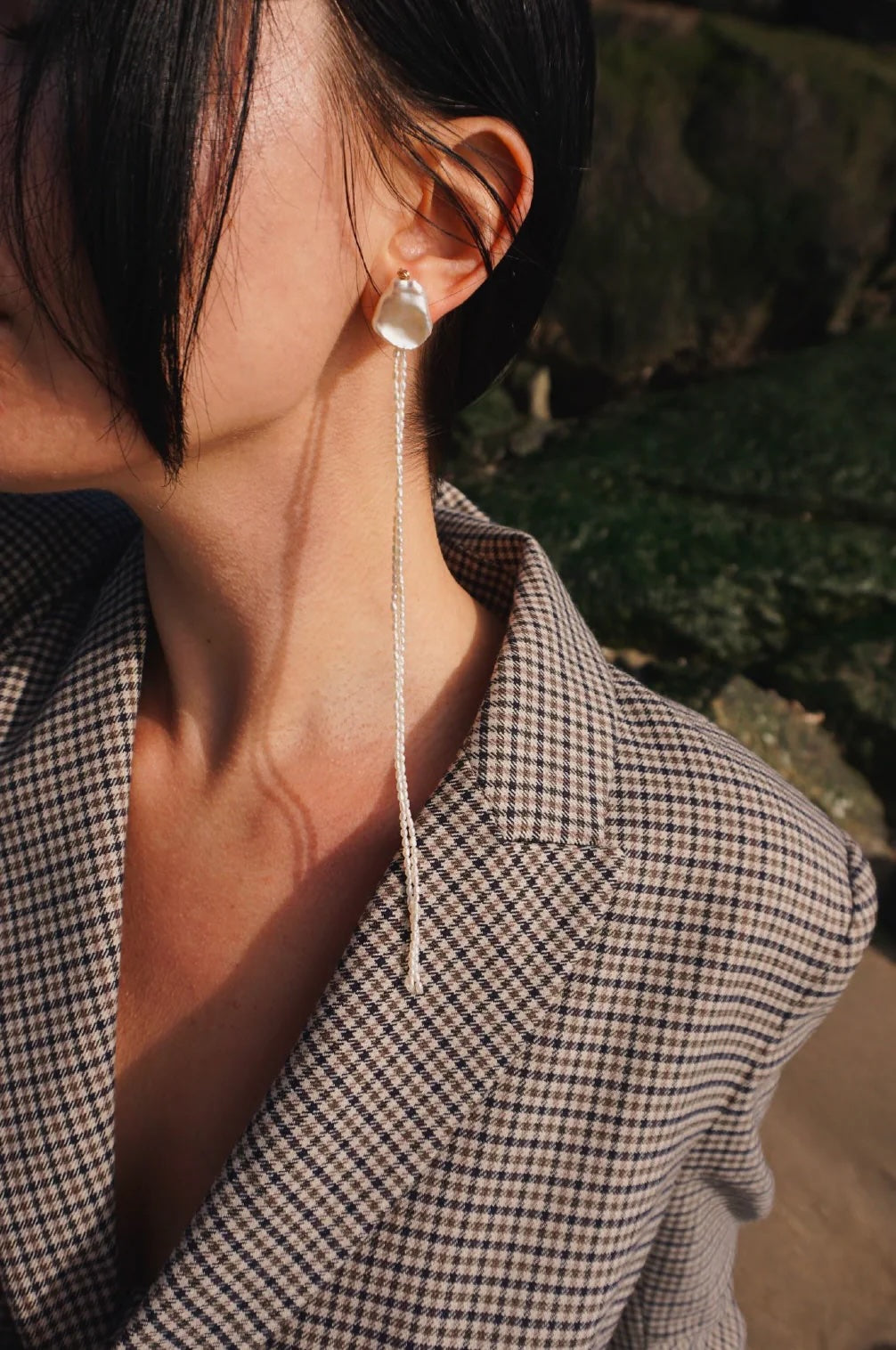 A close-up side profile of a woman wearing a long, dangling pearl earring with a long double strand of pearl bead, paired with a brown and beige checkered blazer.