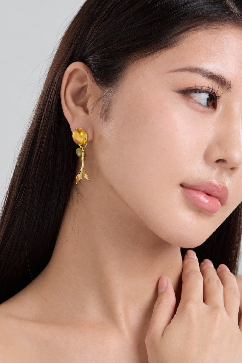 A close-up of a woman wearing a dangling gold earring with a yellow flower and green leaf detail against a light gray background.