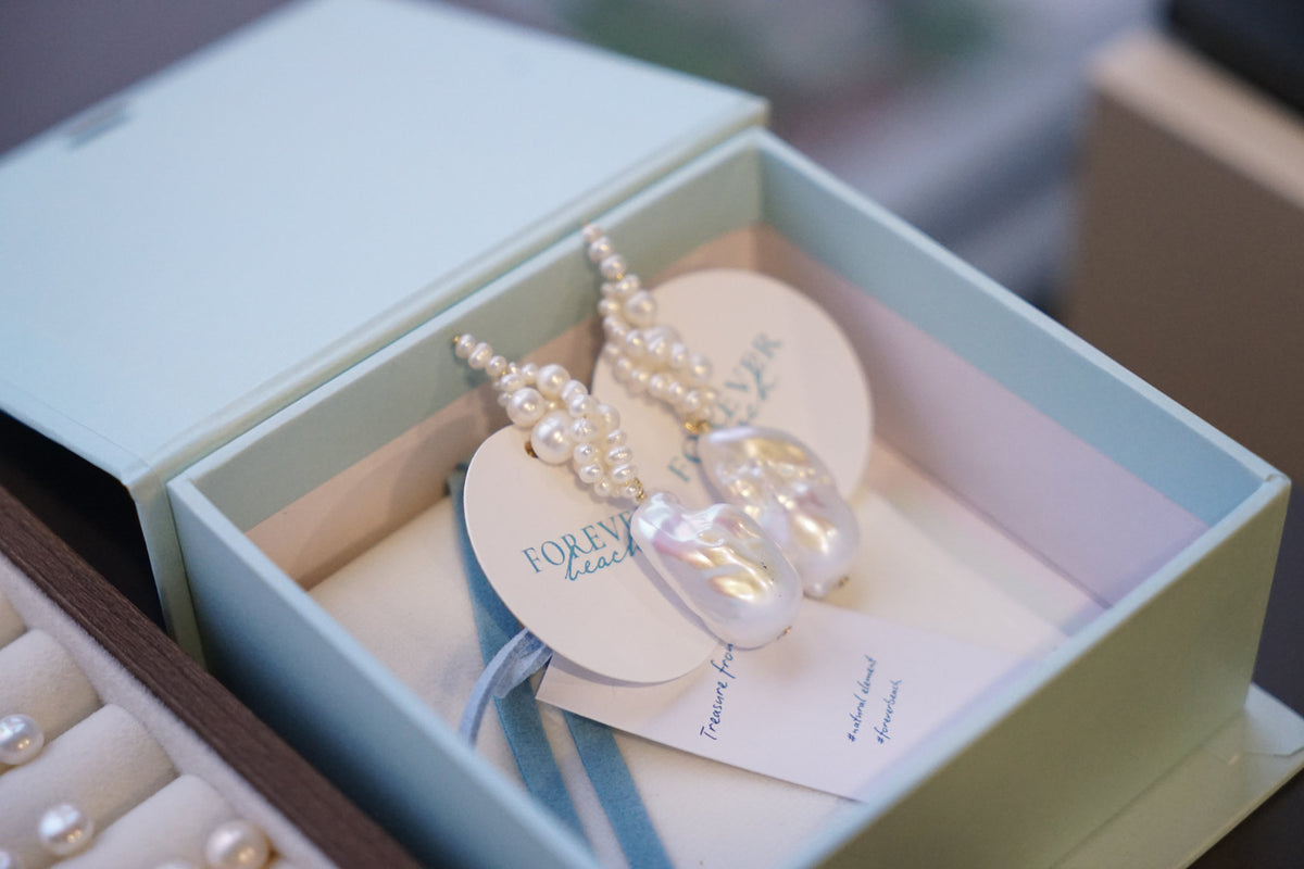 A pair of pearl drop earrings displayed inside a light blue jewelry box with branded tags.