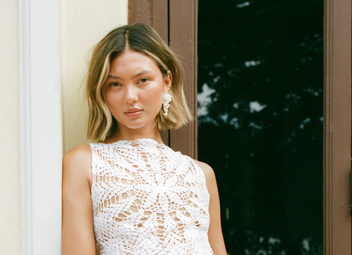 A woman with shoulder-length blonde hair wearing a white sleeveless crochet lace top and a dangling earring made of pearls and a shell, standing next to a dark window frame.
