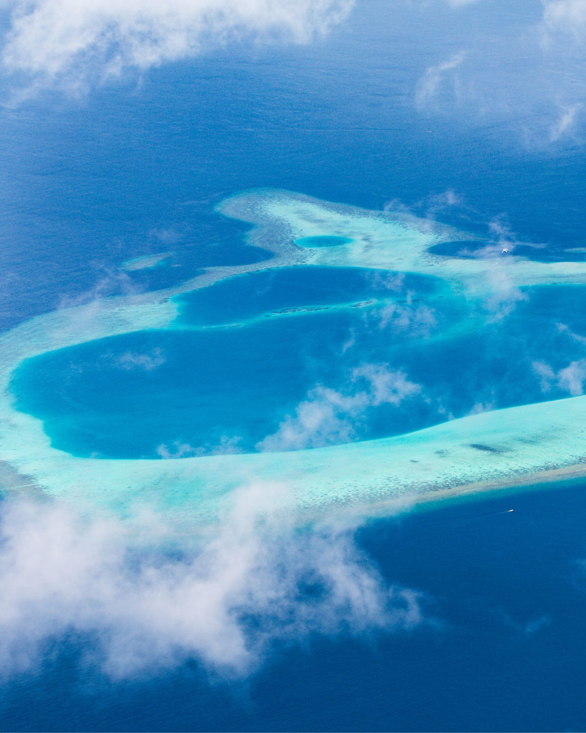 Aerial view of a Maldivian coral atoll with turquoise and deep blue ocean water, white sand, and scattered clouds.
