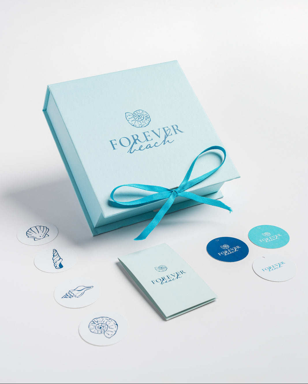 A light blue Forever Beach gift box tied with a turquoise ribbon, shown on a clean white background with matching branded cards, and various small brand tags.