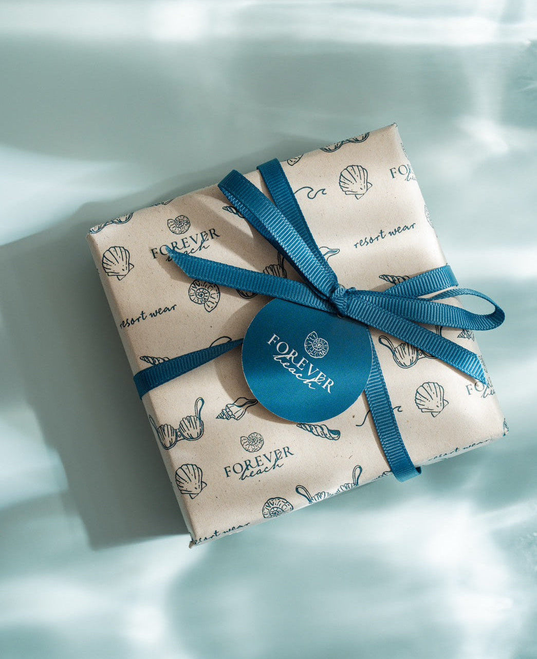 A gift box wrapped in beige paper with a blue seashell pattern and tied with a blue ribbon, featuring a round blue tag that reads 'FOREVER beach' on a soft, light blue background.