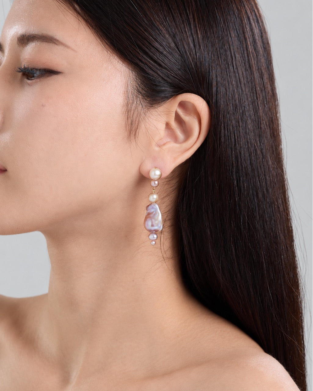 A close-up side profile of a woman wearing a dangling earring. The earring features a small gold-toned bead, a white pearl, and a larger irregular lavender-purple pearls with smaller pearl beads below, set against a neutral gray background.