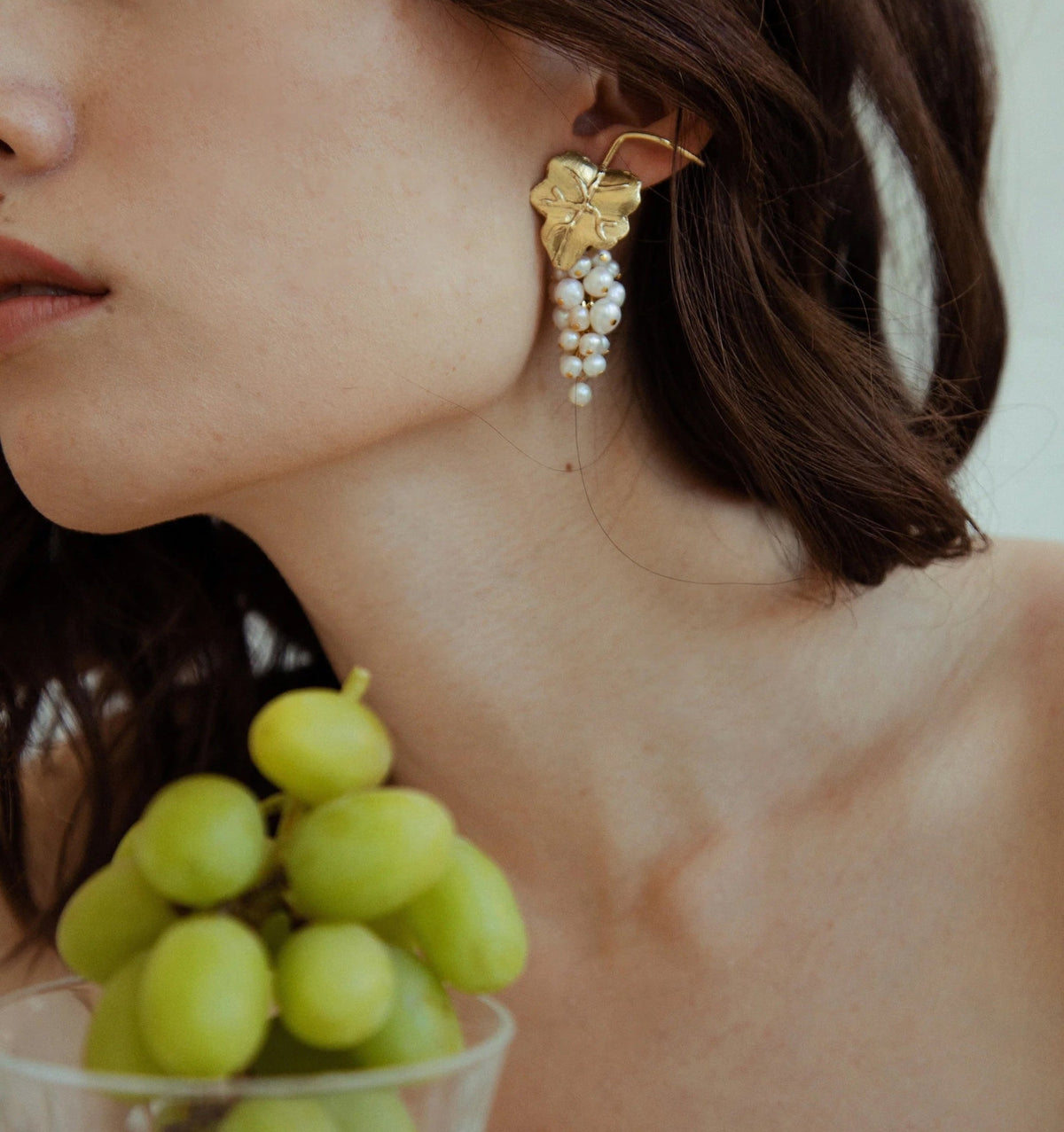 A close-up lifestyle shot of a model wearing a gold leaf-shaped dangle earring adorned with white pearl beads. The model has dark brown hair and is shown from the side, with a small glass bowl of green grapes in the foreground.