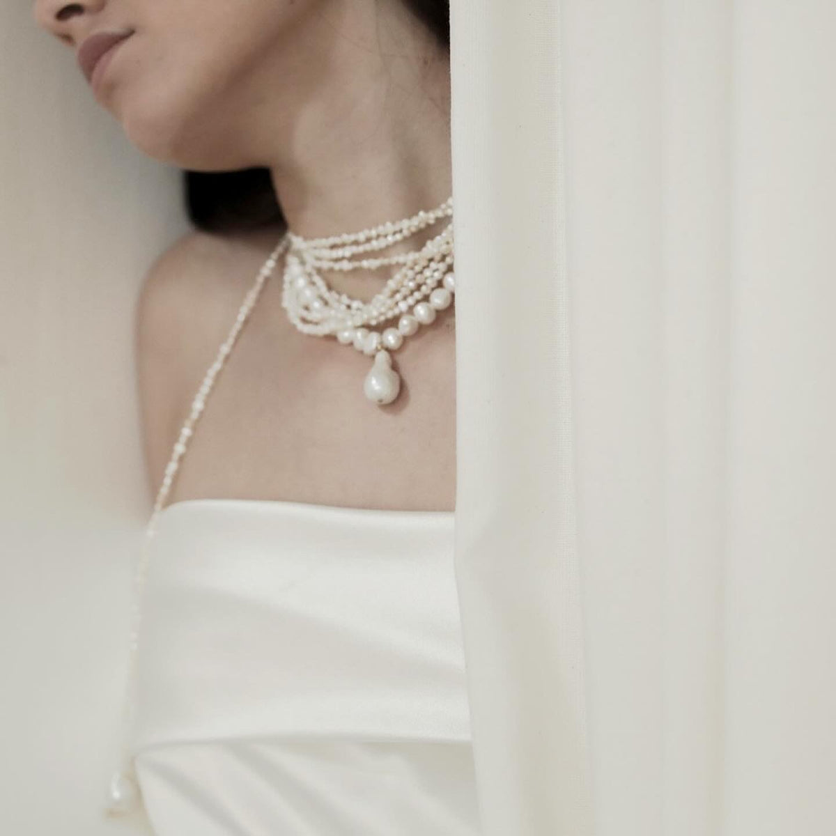 A close-up of a model wearing a multi-strand pearl necklace with a teardrop pearl pendant, styled with a white strapless dress and a sheer white curtain.