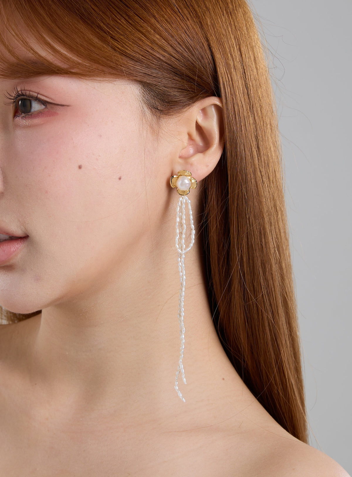A close-up of a woman with long auburn hair wearing a long dangle earring. The earring features a gold-toned setting with a white pearl at the top and cascading strands of small white beads.