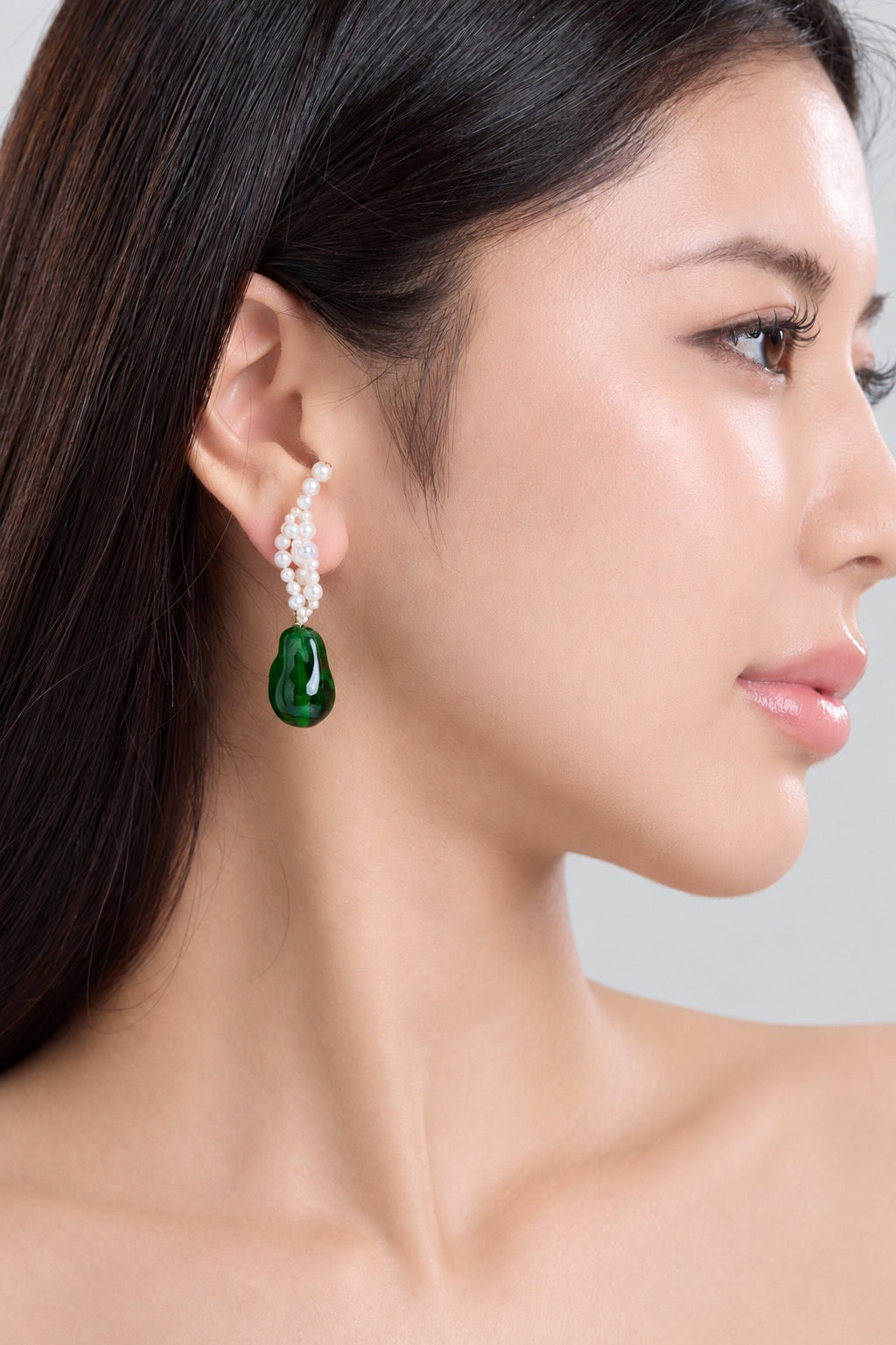 A close-up side profile of a woman wearing a dangling earring with a green teardrop-shaped gemstone and a curved top made of small white pearls, against a light gray background.