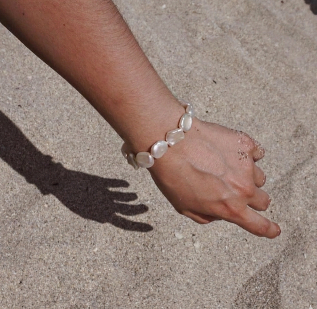 A close-up of a person's wrist and hand wearing a bracelet made of irregular white keshi pearls, set against a sandy beige background with a distinct hand shadow.