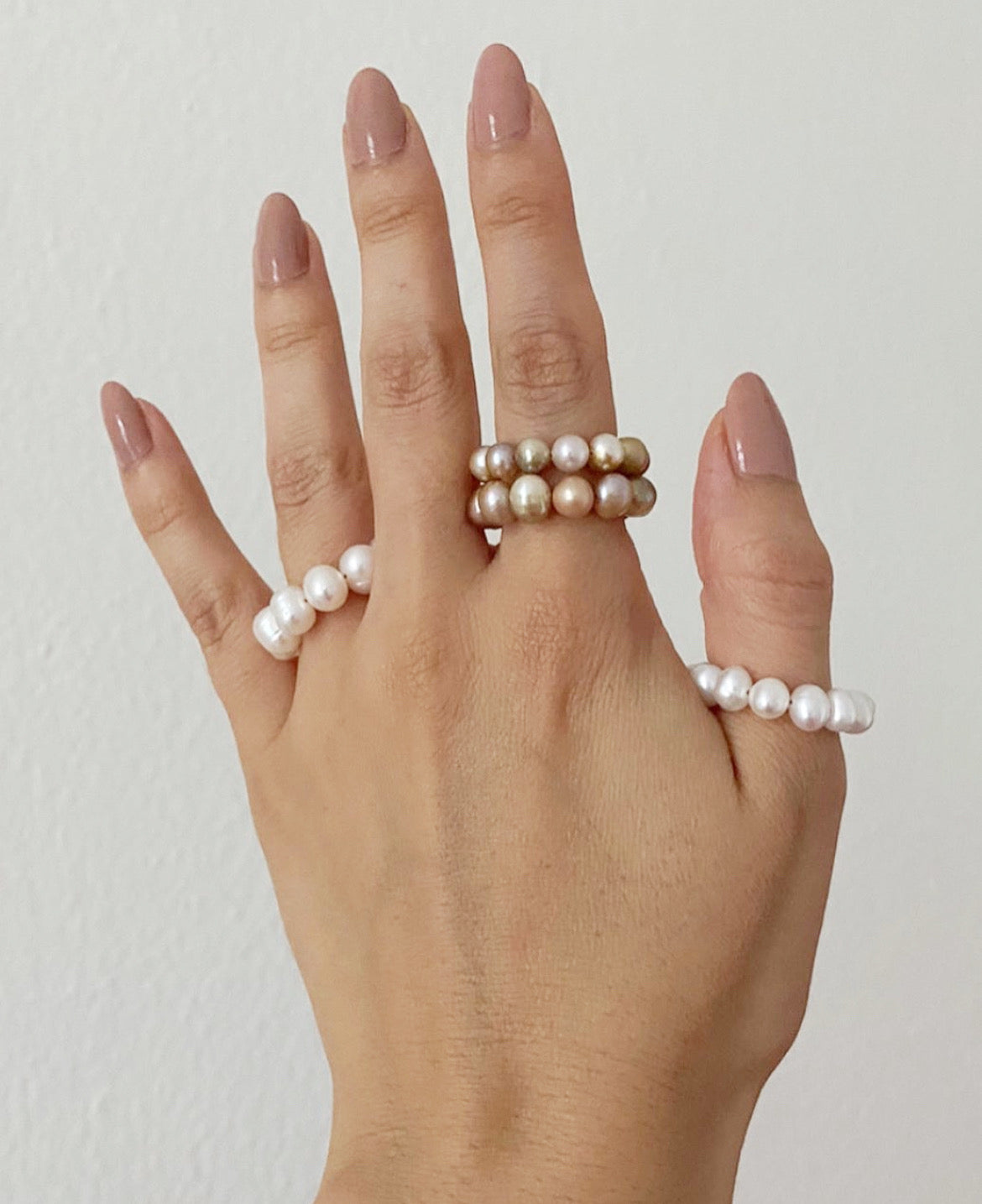 A hand with manicured nails wearing multiple pearl rings, including a double-row pearl ring on the middle finger, a single pearl ring on the ring finger, and a single pearl ring on the thumb, set against a plain off-white wall.