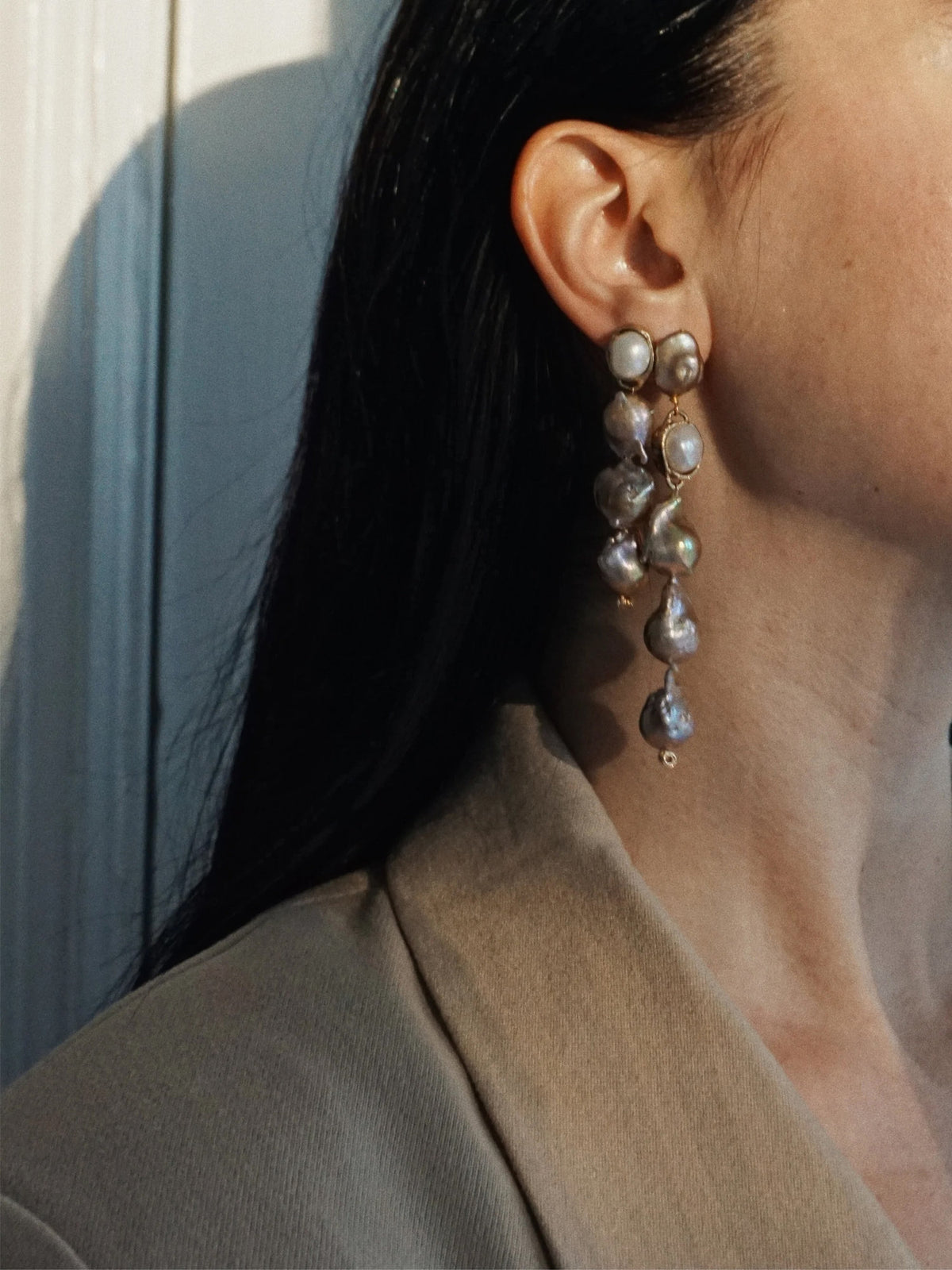 A close-up side profile of a woman wearing a long, dangling drop earring made of irregular baroque pearls in a gold-toned setting, paired with a beige blazer.