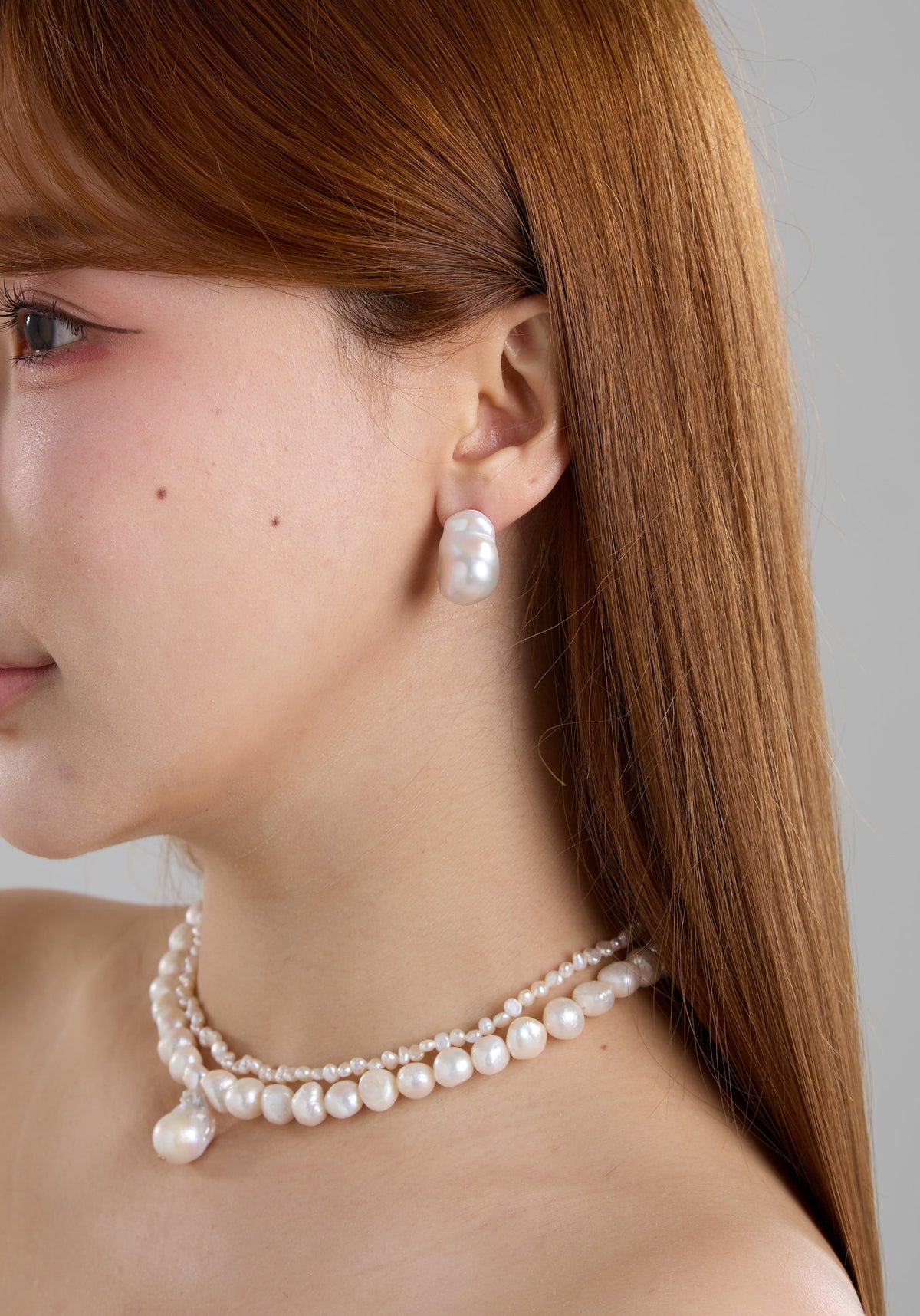 A close-up side profile of a woman with long auburn hair, wearing a double-strand pearl necklace and a matching pearl drop earring against a neutral gray background.