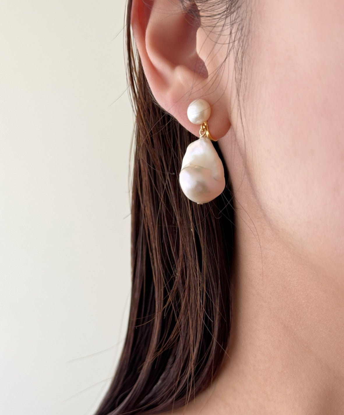 A close-up of a model's ear wearing a double-drop dangle earring. The earring features a small white pearl stud on the top drop and a larger, irregular baroque-style white pearl drop on the bottom drop, connected by a small gold-toned metal link.