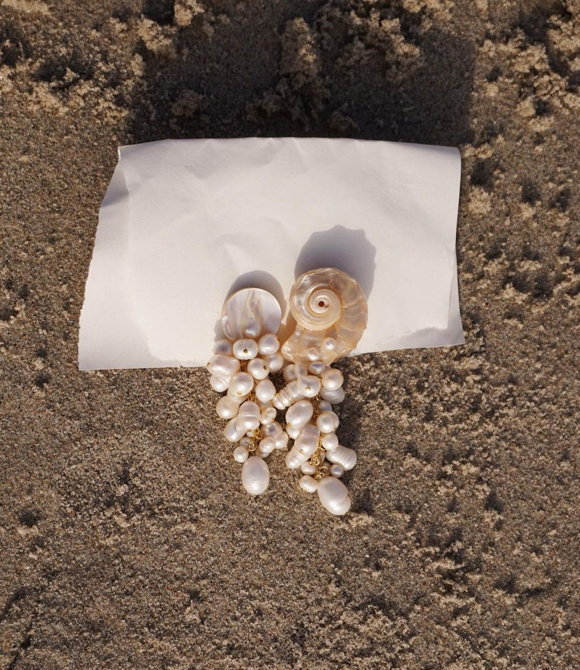 A pair of shell and pearl drop earrings resting on a small white piece of paper placed on sandy ground.