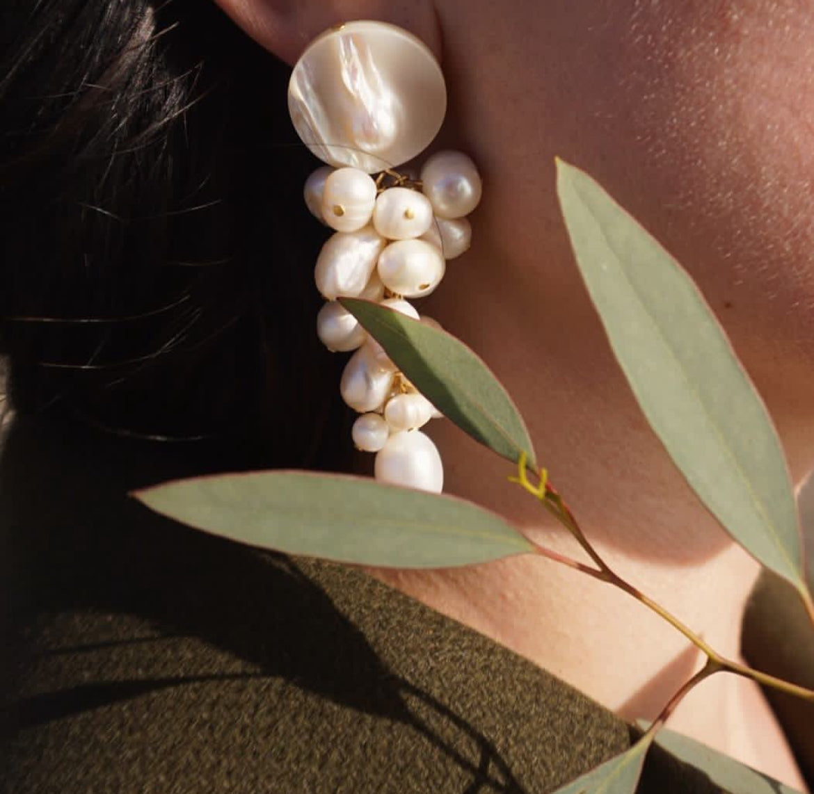 A close-up of a person wearing a long, multi-tiered dangle earring made of large and small white pearls with a gold-toned accent, next to a leafy branch.