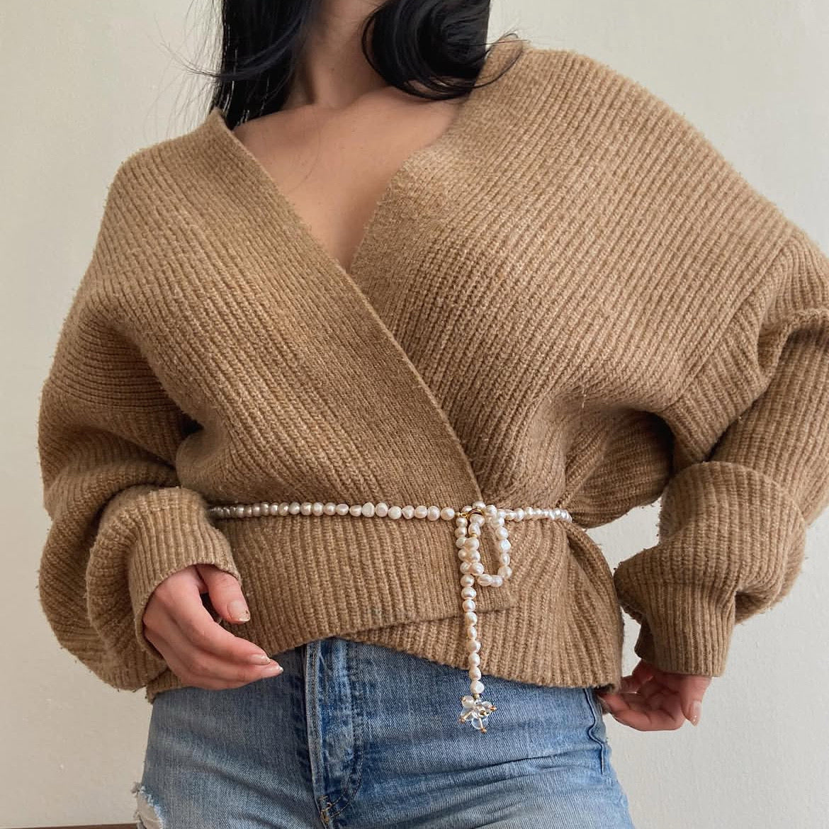 A person wearing a pearl beaded belt with a small decorative charm made of quartz gem at the end. The person is wearing a sweater in camel brown, and blue jeans.